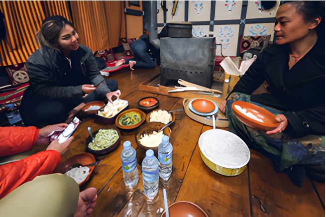 Guests sharing a traditional home-cooked Bhutanese dinner inside a local farmhouse, seated on the floor around wooden bowls of rice and dishes, reflecting warm hospitality and everyday rural life in Bhutan.